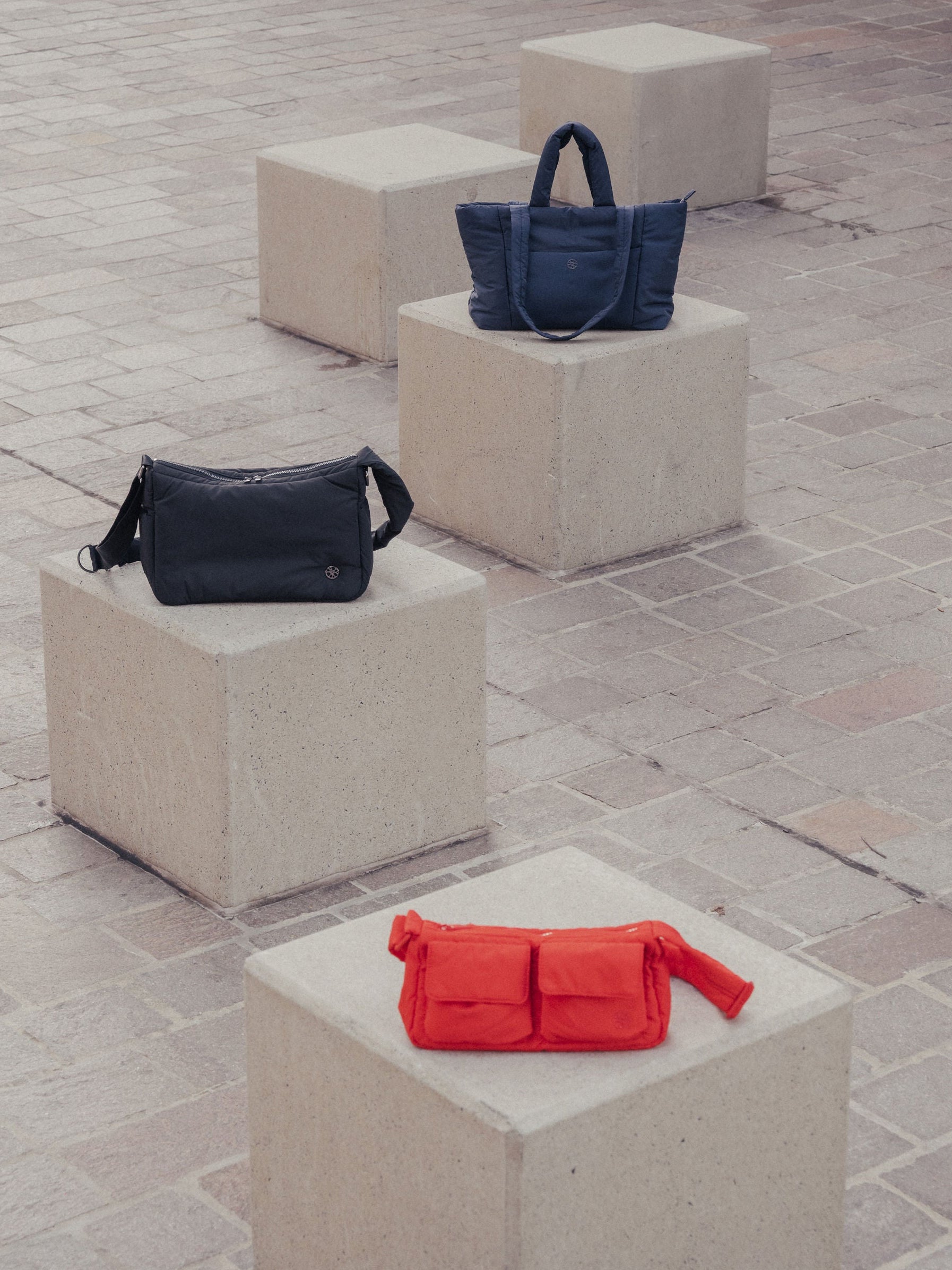Three bags, one black, one blue and one red, on concrete cubes with a stone pavement background.